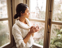 A young woman in a sweater holds a mug and looks out a window.
